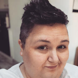 Headshot of Lola Keeley, author. A white cis woman in her late thirties with short dark hair in a faux hawk style. She is looking side on at the camera and smiling slightly.